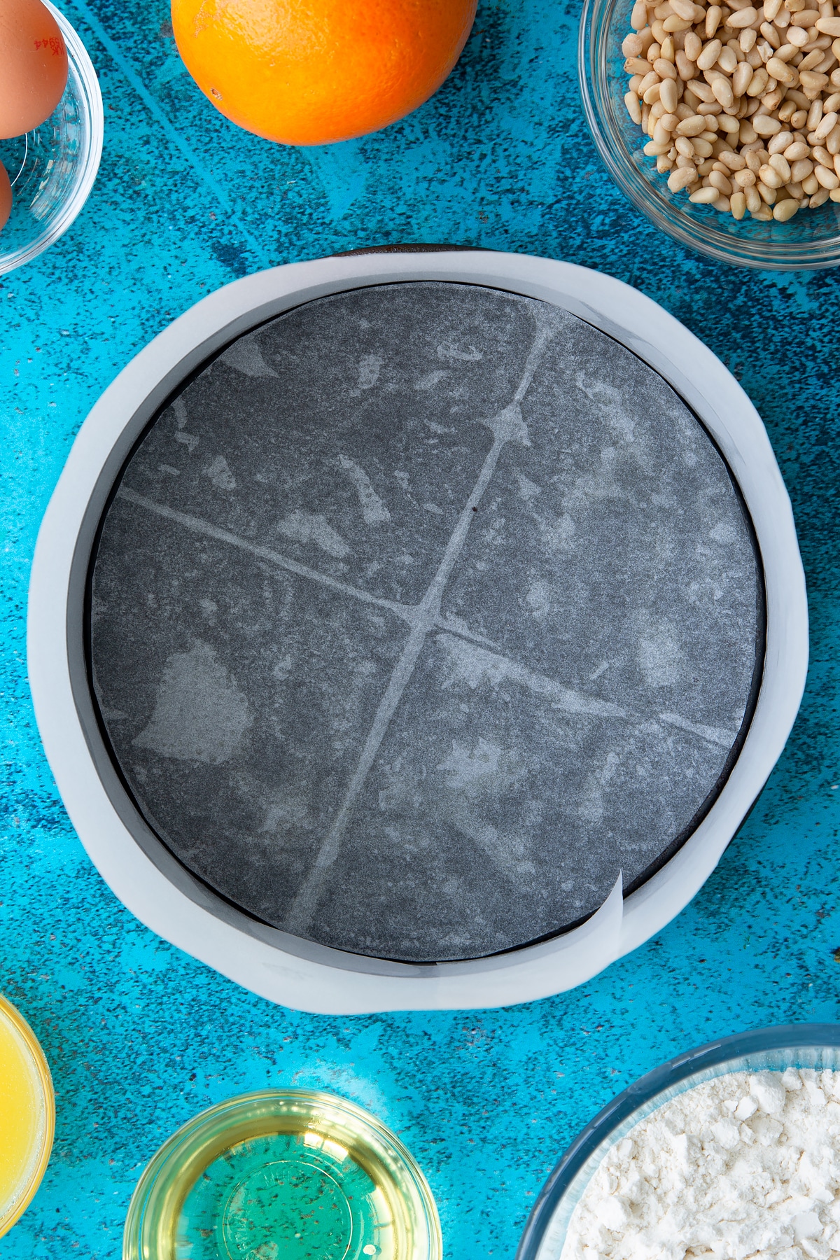 A greased and lined cake tin. Ingredients to make pine nut cake surround the bowl.