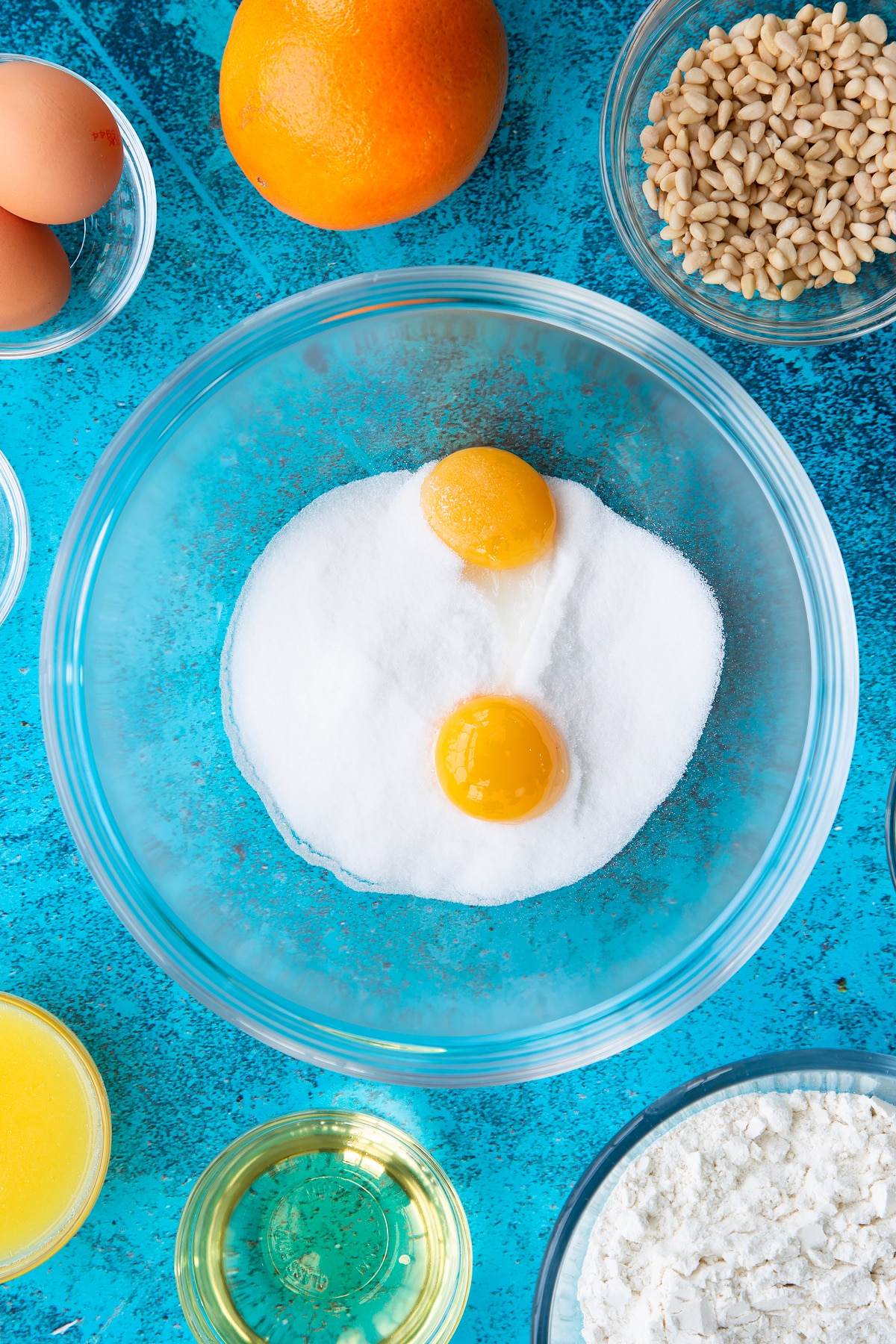 Sugar and egg yolks in a bowl. Ingredients to make pine nut cake surround the bowl.