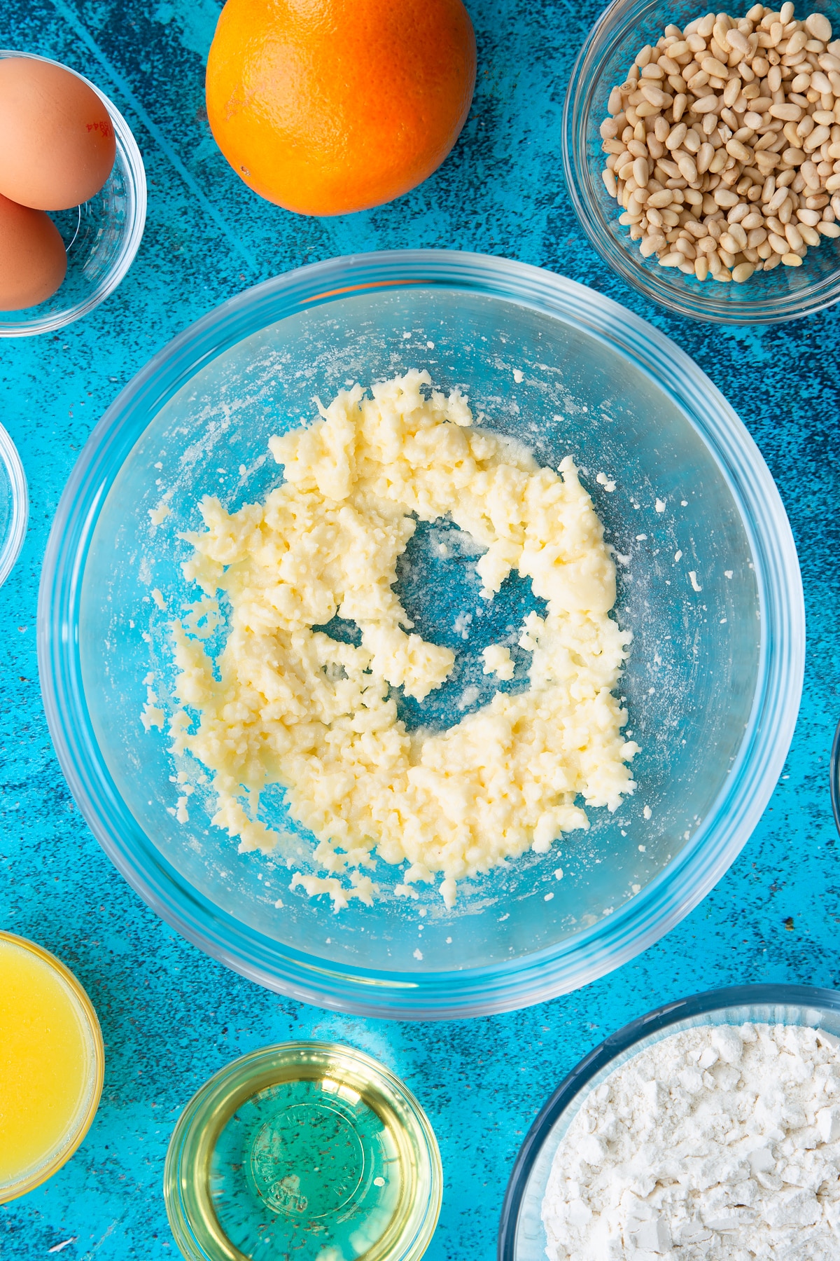 Sugar and egg yolks whisked together in a bowl. Ingredients to make pine nut cake surround the bowl.