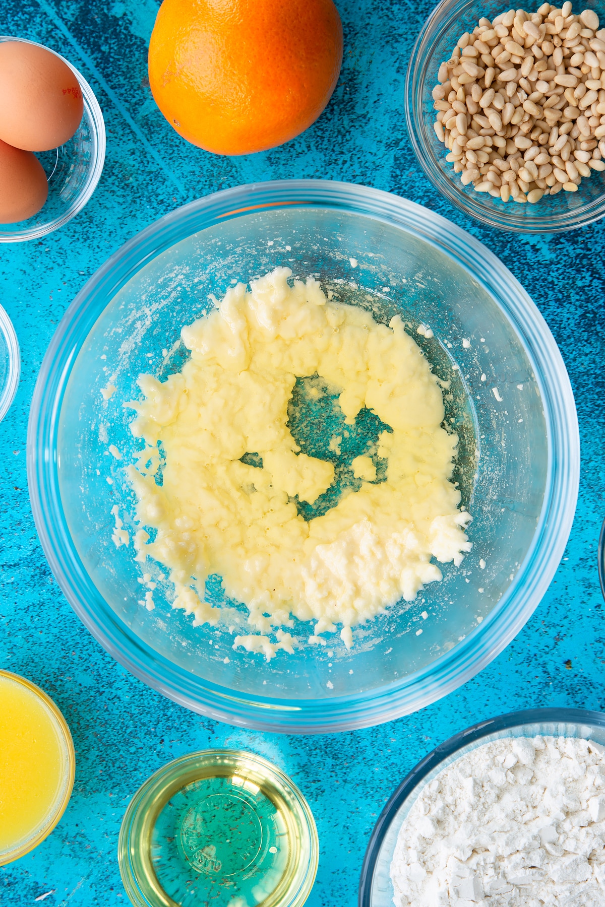 Sugar and egg yolks whisked together in a bowl with oil on top. Ingredients to make pine nut cake surround the bowl.