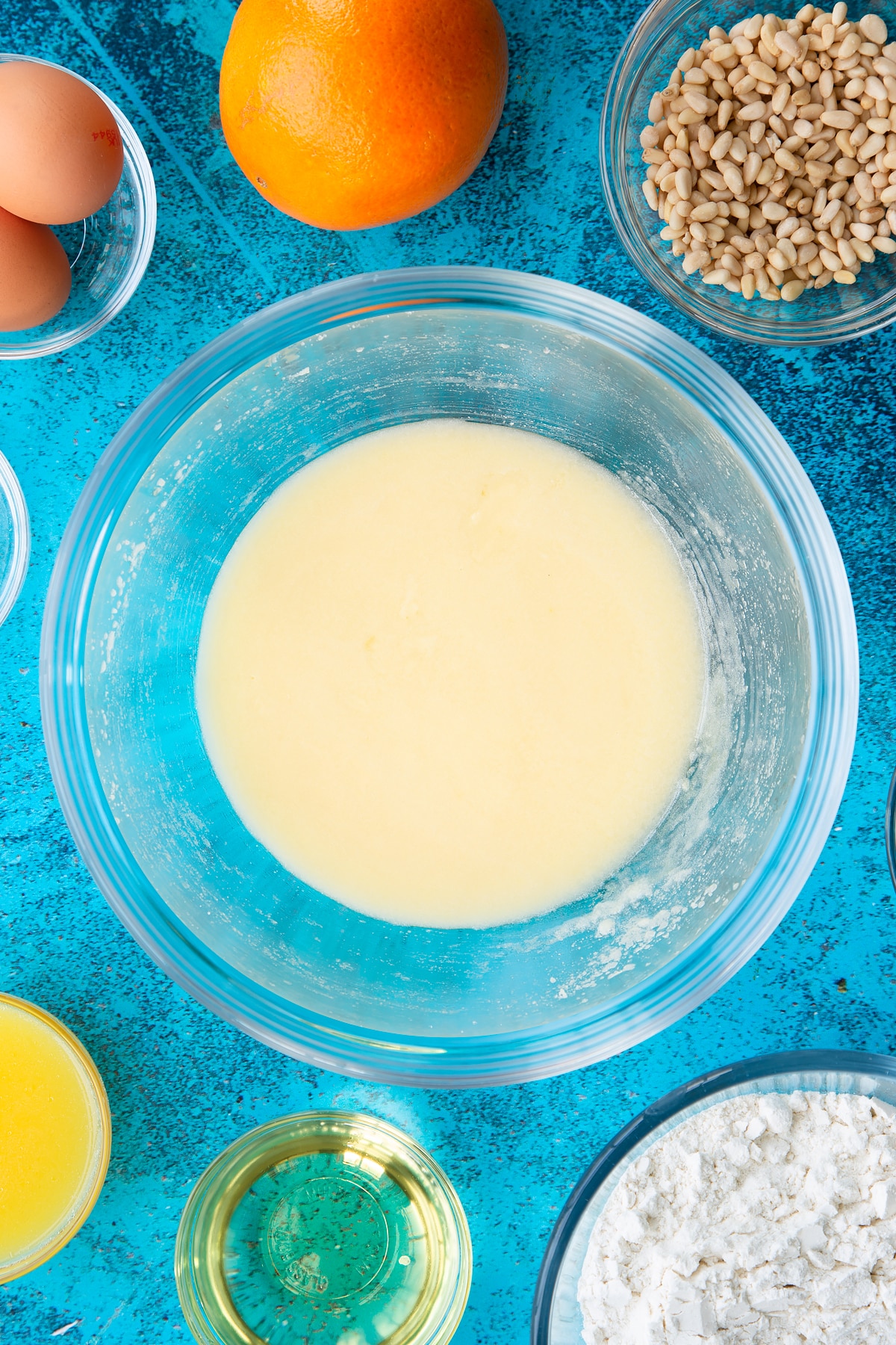 Sugar, egg yolks and oil whisked together in a bowl. Ingredients to make pine nut cake surround the bowl.