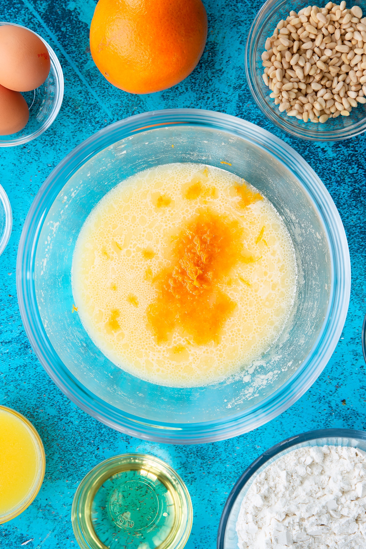 Sugar, egg yolks and oil whisked together in a bowl with orange juice and zest on top. Ingredients to make pine nut cake surround the bowl.
