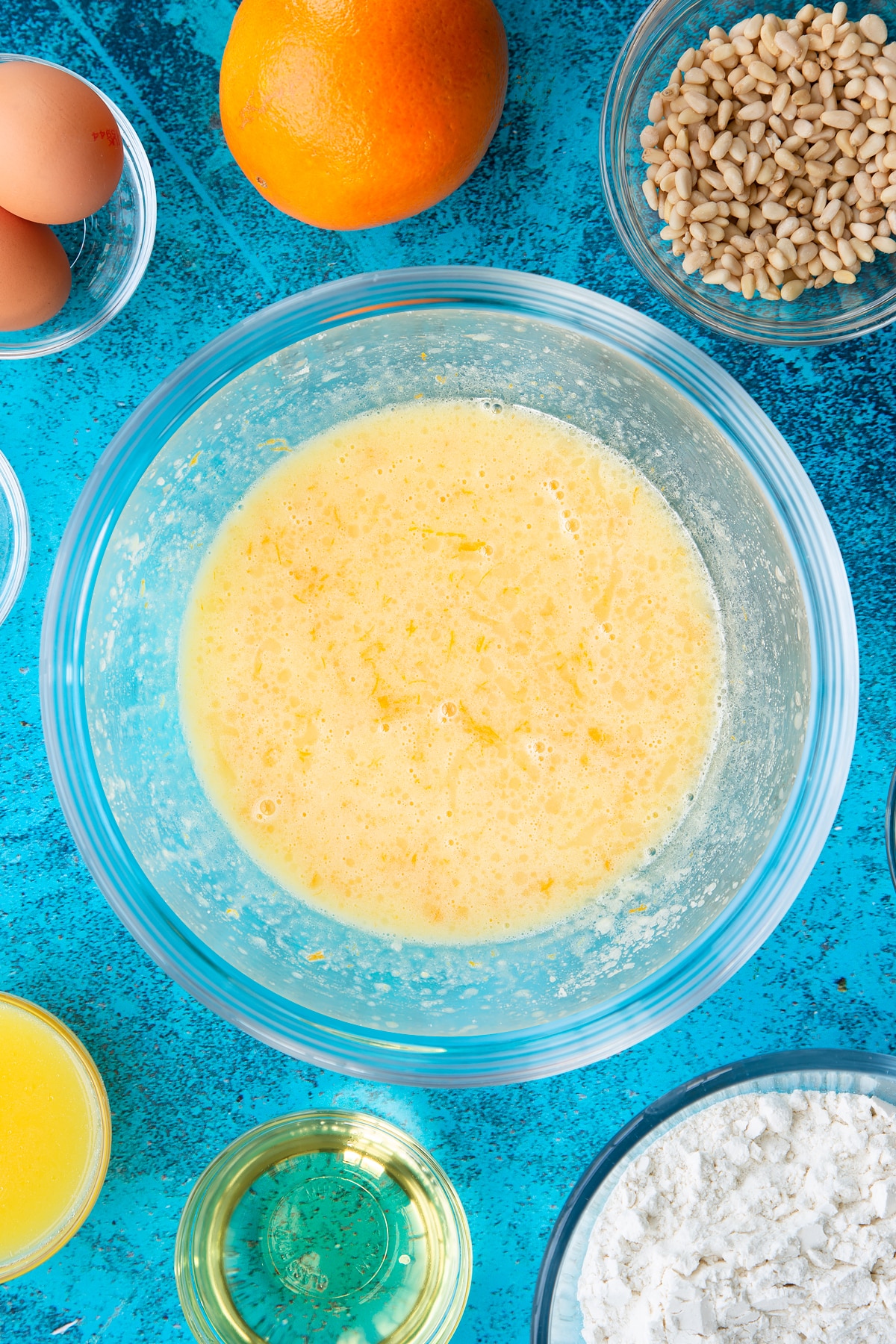 Sugar, egg yolks, oil, orange juice and zest whisked together in a bowl. Ingredients to make pine nut cake surround the bowl.