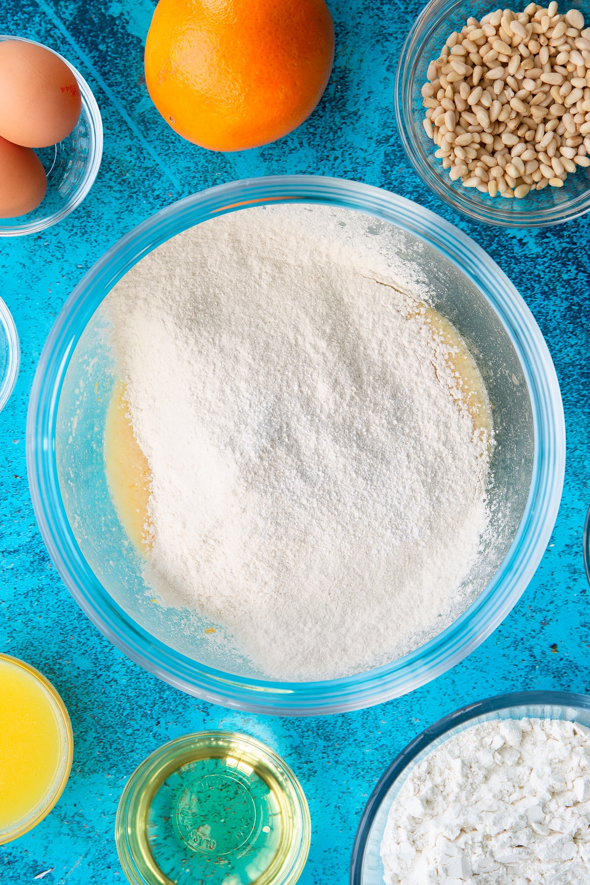 Sugar, egg yolks, oil, orange juice and zest whisked together in a bowl with flour and baking powder on top. Ingredients to make pine nut cake surround the bowl.