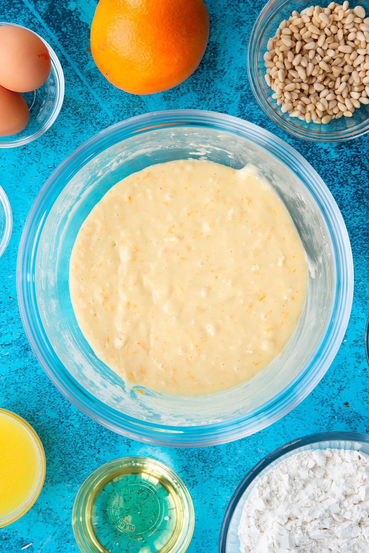 Sugar, egg yolks, oil, orange juice, zest, flour and baking powder whisked together in a bowl. Ingredients to make pine nut cake surround the bowl.