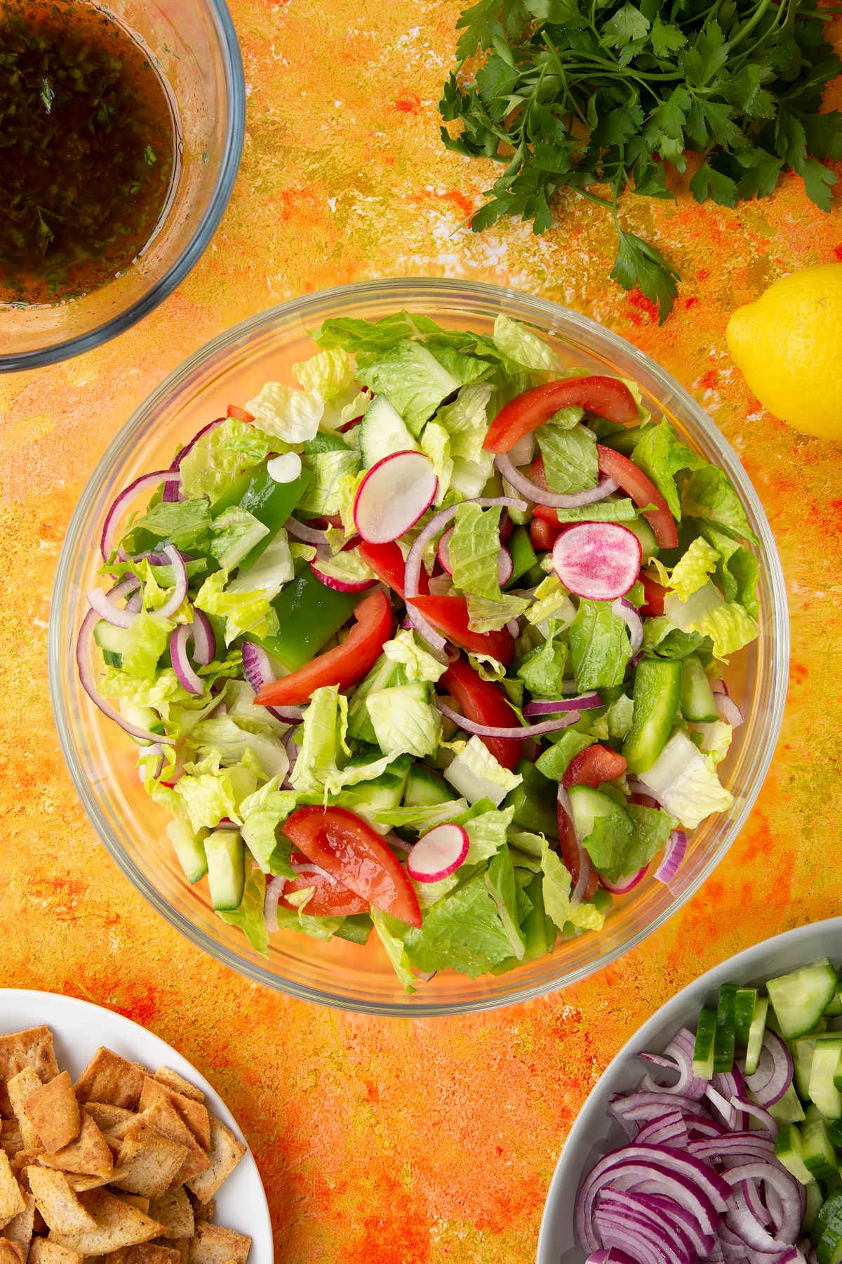 Lettuce, tomatoes, red onion, cucumber, green peppers and radish mixed together in a glass mixing bowl. Ingredients to make vegan fattoush surround the bowl.