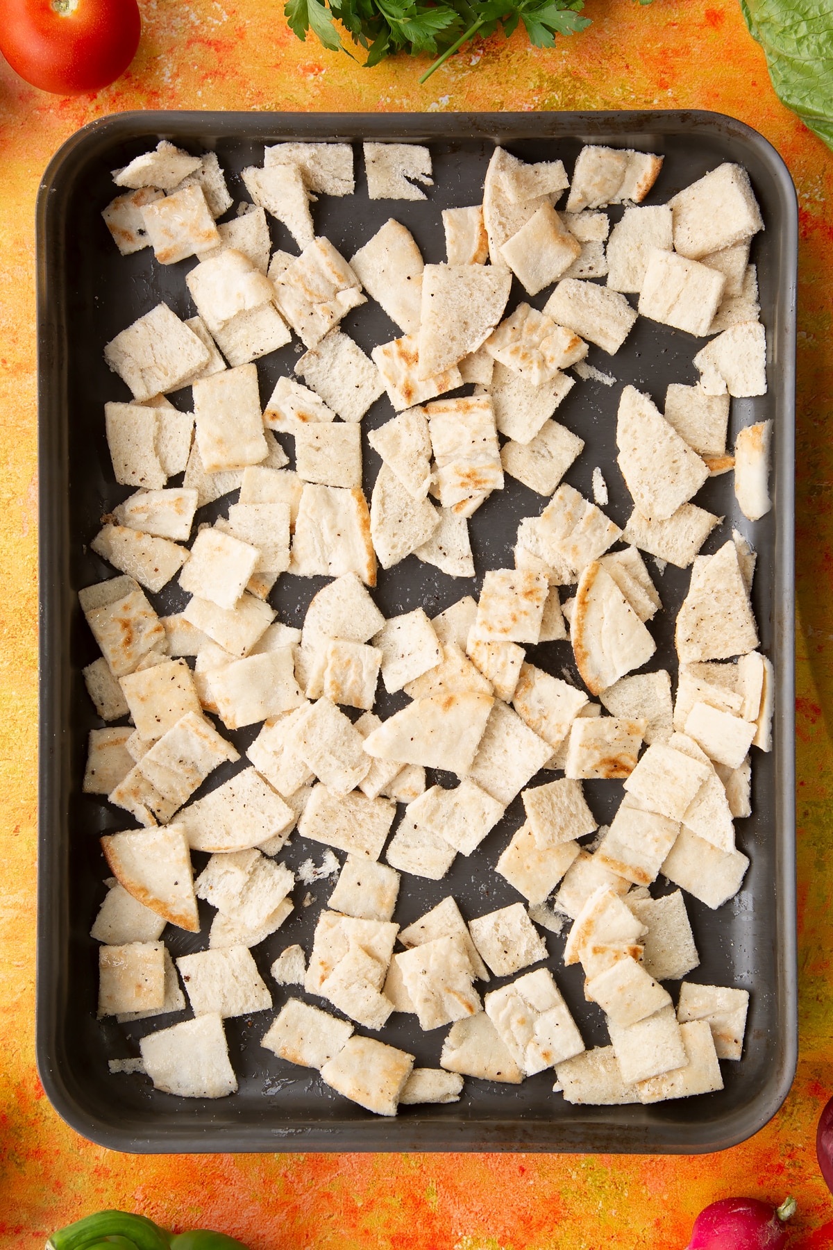 Pitta pieces in a tray, seasoned with olive, salt and pepper. Ingredients to make vegan fattoush surround the tray.