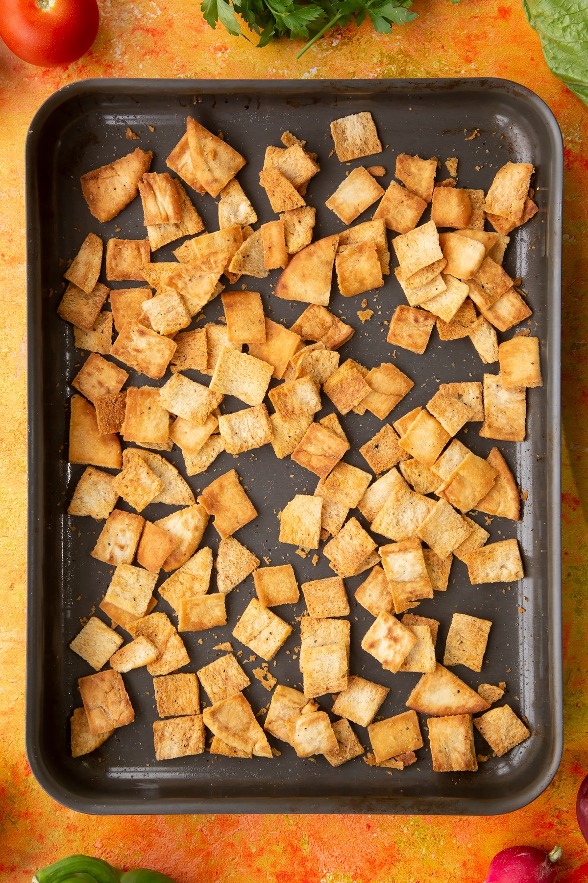 Baked pitta pieces in a tray. Ingredients to make vegan fattoush surround the tray.