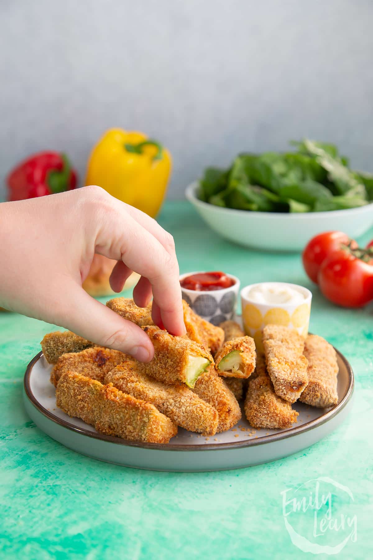 Hand reaching in to grab half of one of the finished baked courgette fries off a decorative plate.