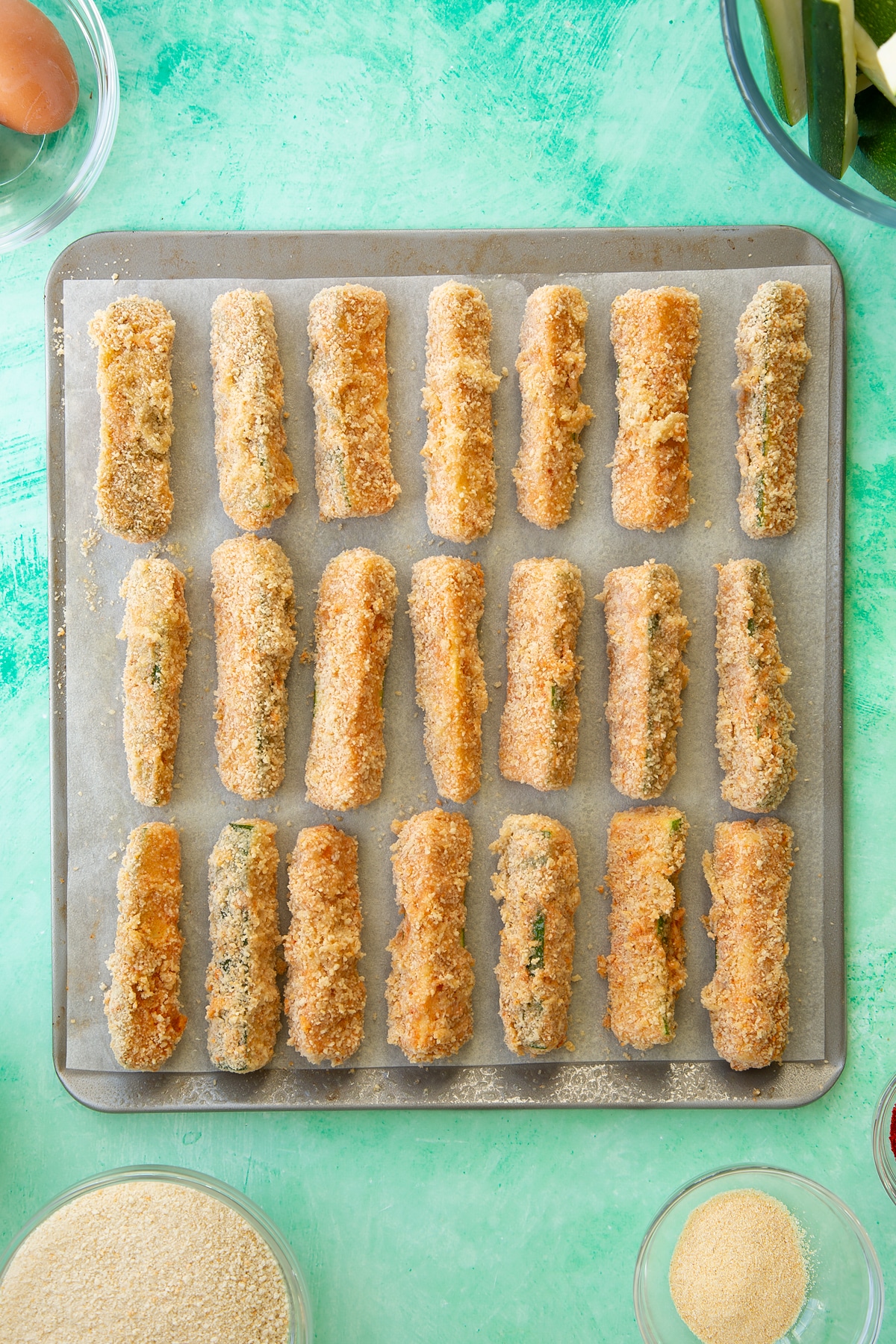 Overhead shot of a baking tray with multiple courgette slices covered in flour, egg and breadcrumbs.