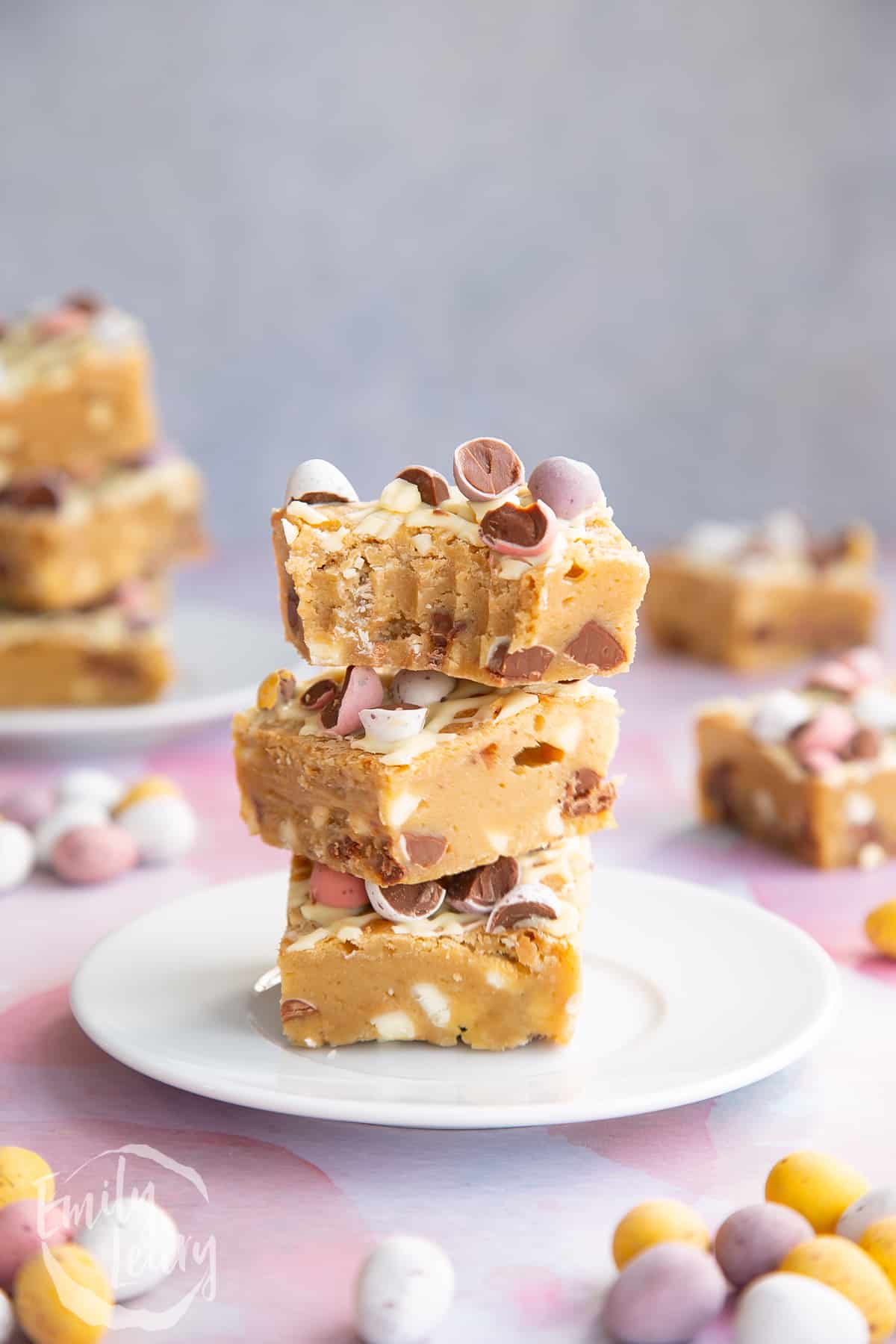 Three Mini Egg blondies on a white plate. More Mini Egg blondies are behind. The top one has been bitten.