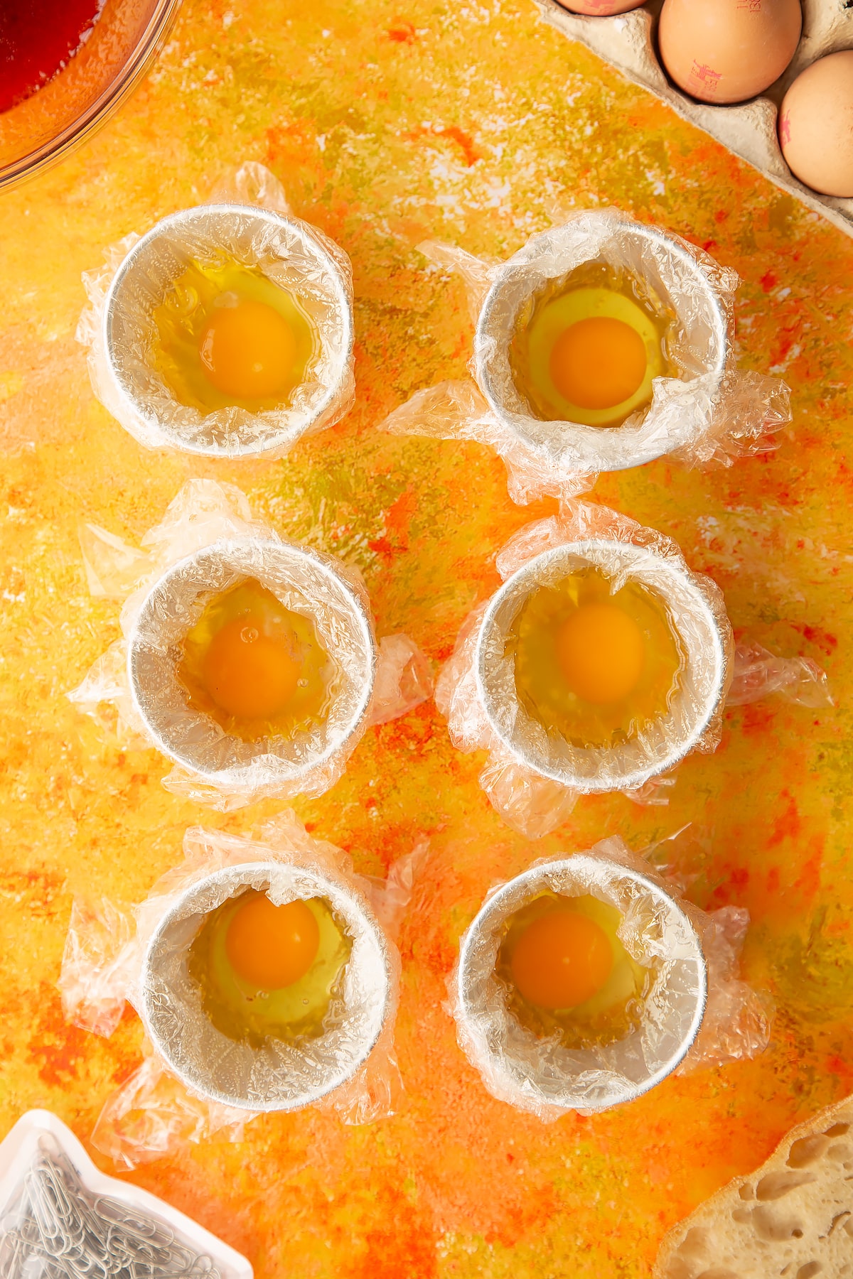 Overhead shot of six small bowls i.e. ramkins covered in clingfilm and oil with an egg cracked inside.