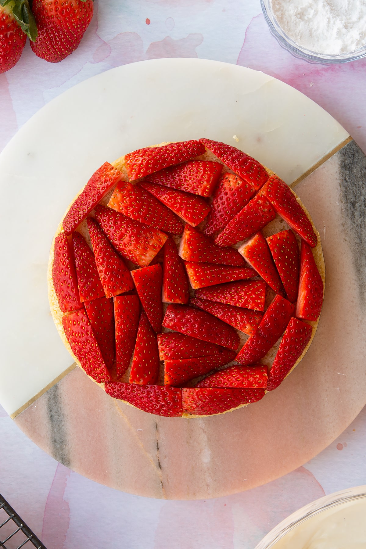 circular sponge cake on a pink marble board topped with chopped strawberries.