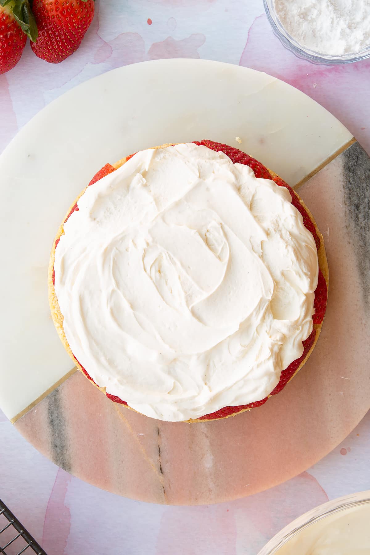 circular sponge cake topped with chopped strawberries and fresh cream on a pink marble board.