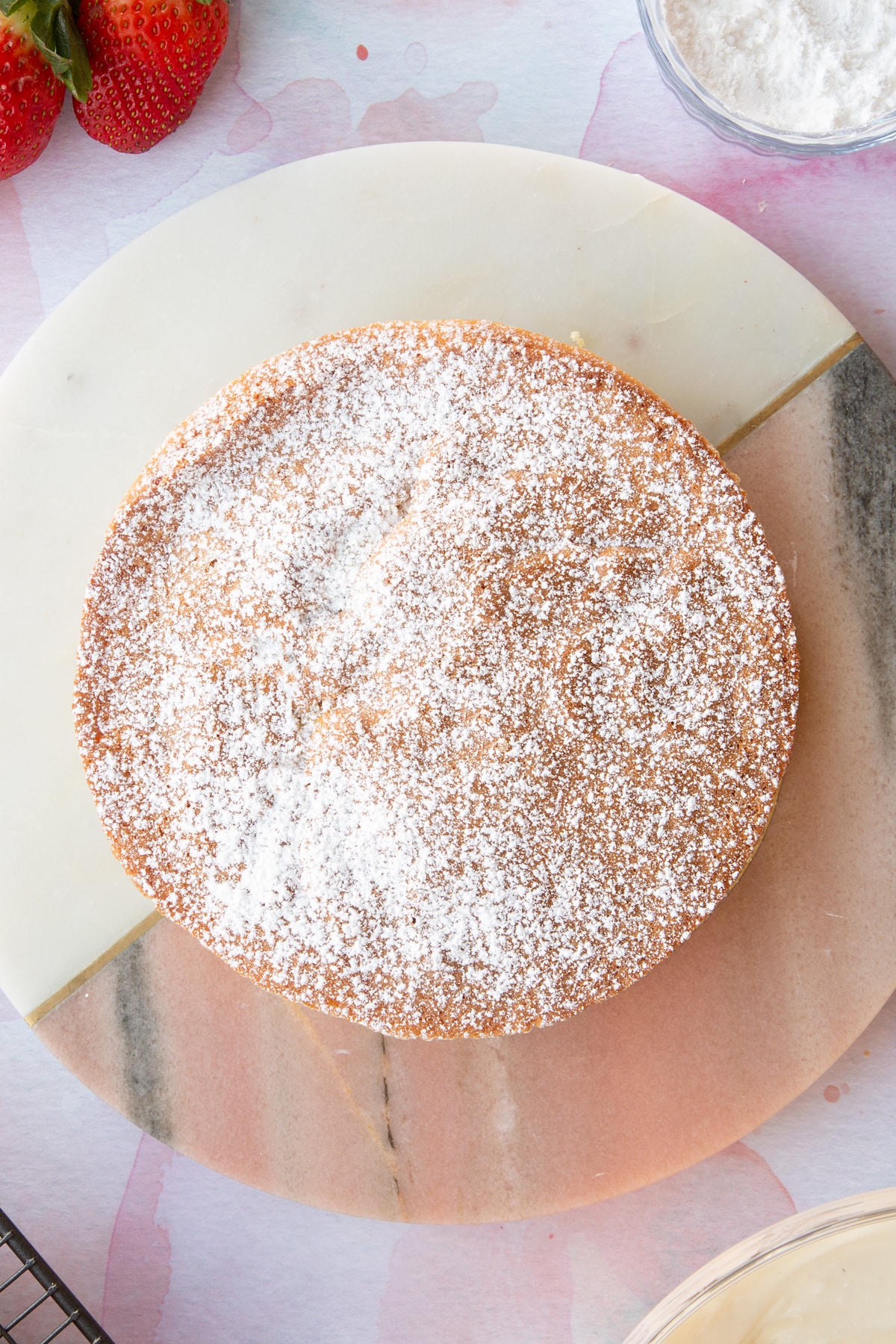 circular strawberry cream sponge cake topped with a dusting og icing sugar on a pink marble board.