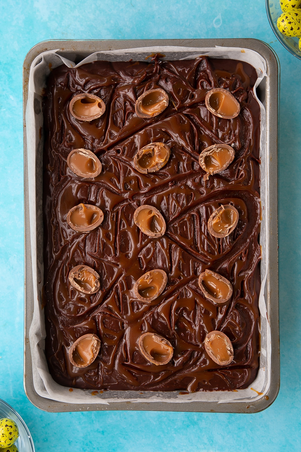 Overhead shot of the halved mini caramel eggs being added to the top of the easter brownies mixture inside the rectangular cake tin.