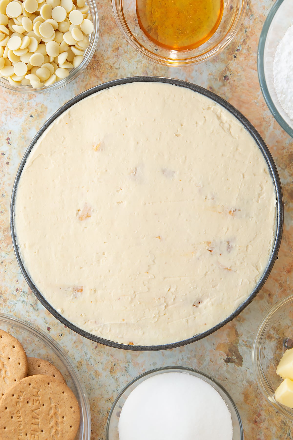 whisked white chocolate cheesecake topping in a large clear bowl topped with honeycomb crumbs pressed into a cake tin.