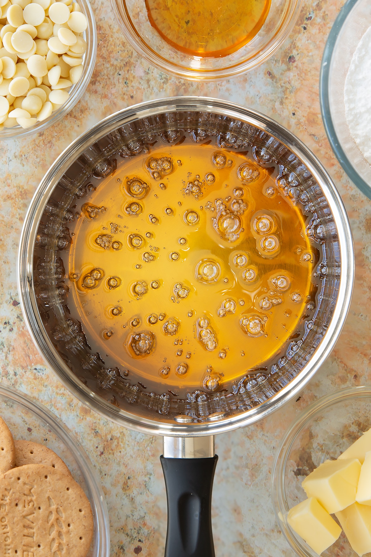 bubbling transparent sugar and water mixture in a saucepan surrounded by ingredients in small clear bowl.