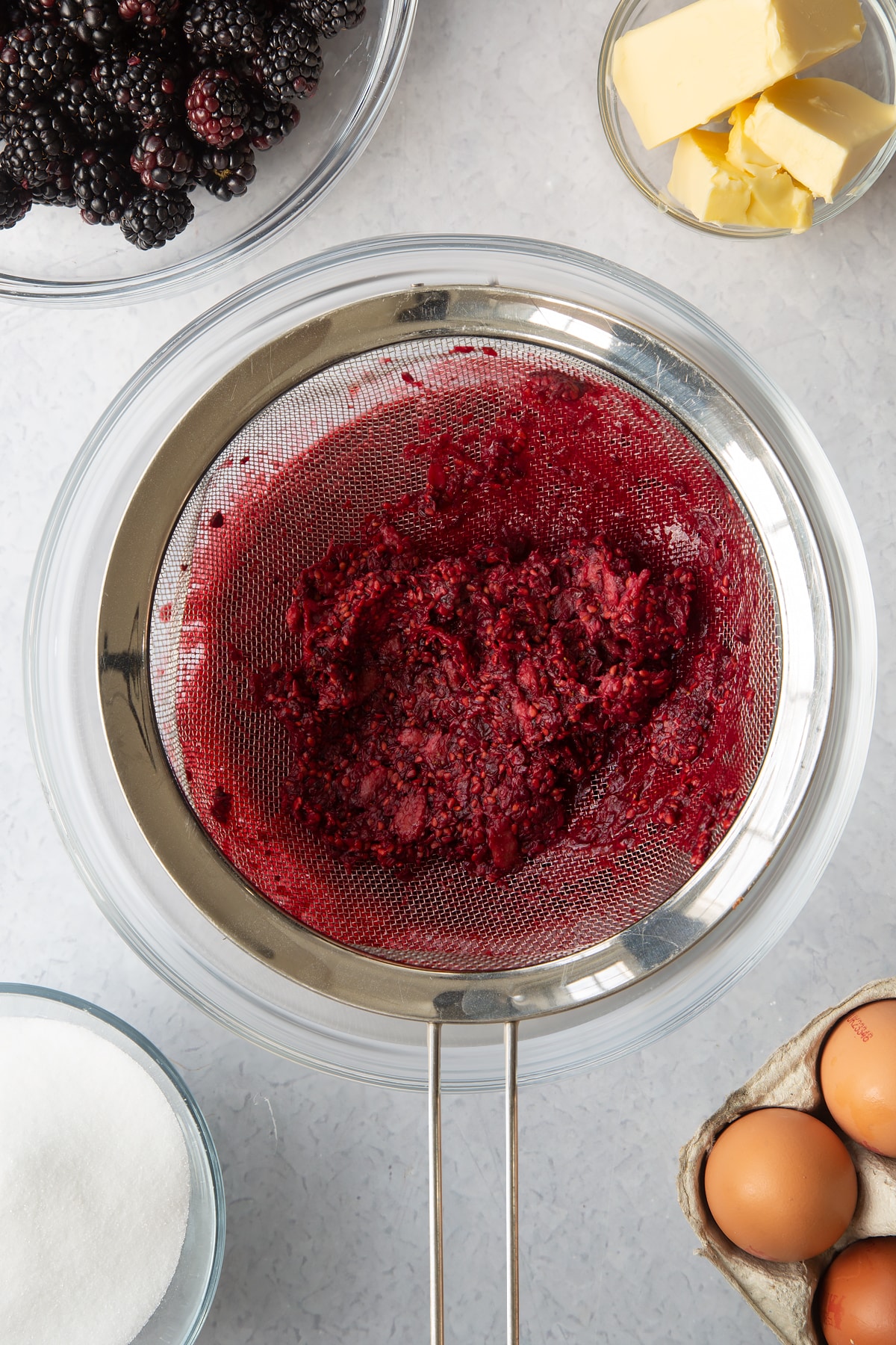 mushed blackberries in a silver sieve over a large clear bowl.