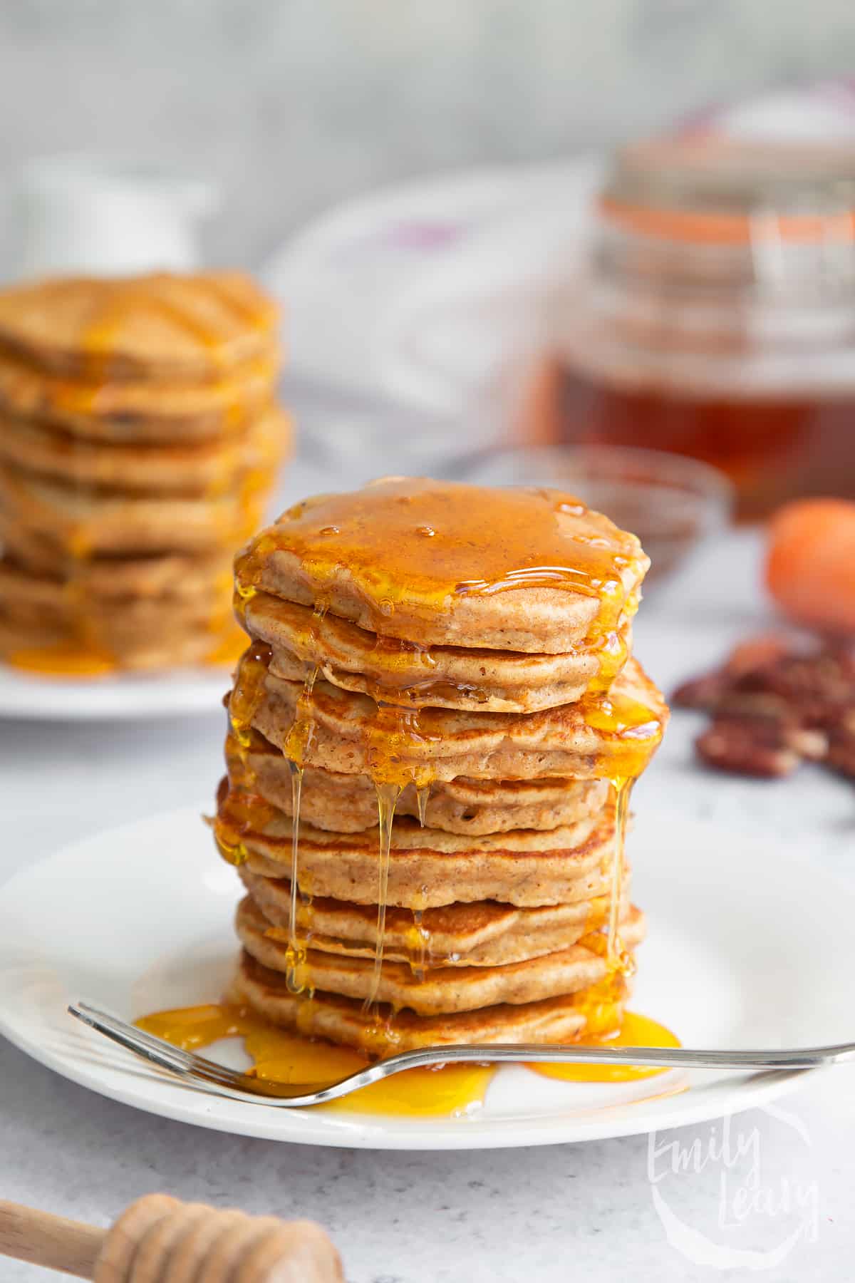 a stack of carrot cake pancakes on a white plare with more pancakes in the background topped with honey.