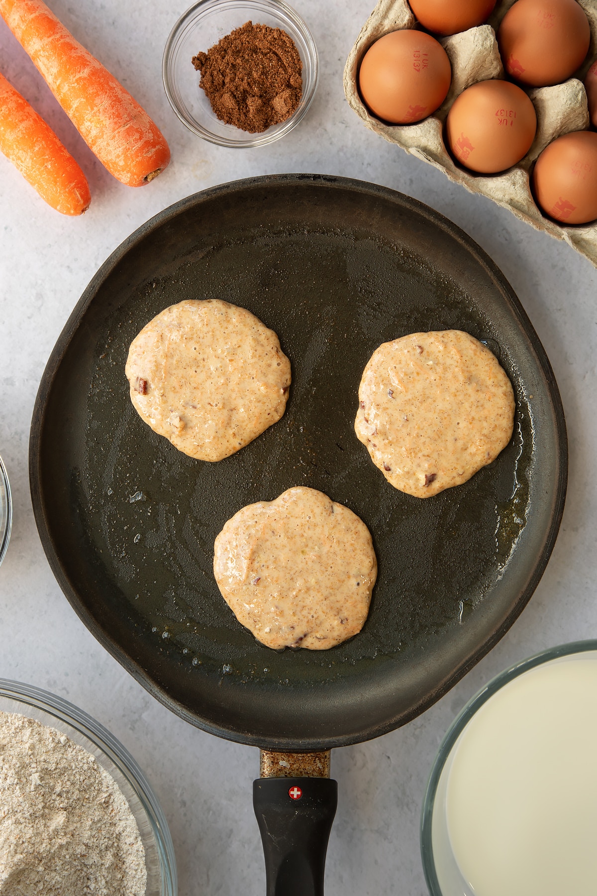 3 carrot cake pancake batter rounds in a large frying pan.