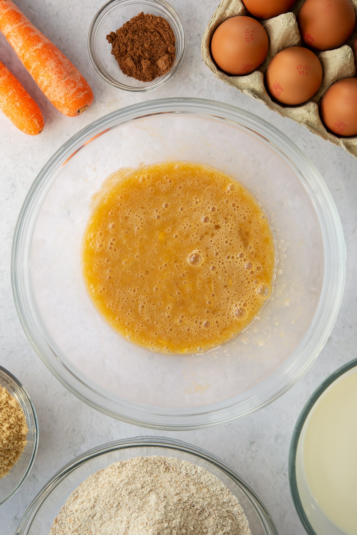 whisked eggs and sugar mixture in a large clear bowl surrounded by ingredients.