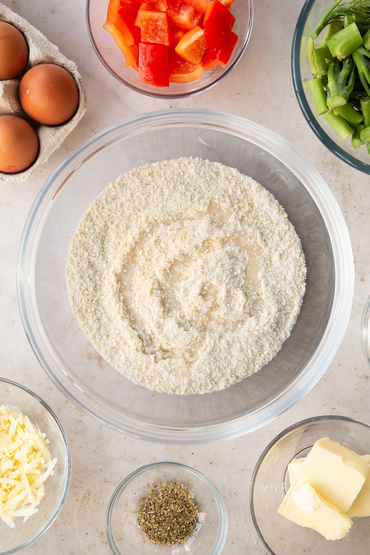 butter and flour crumbed mixture in a large clear mixing bowl white water drizzled on top surrounded by ingredients.