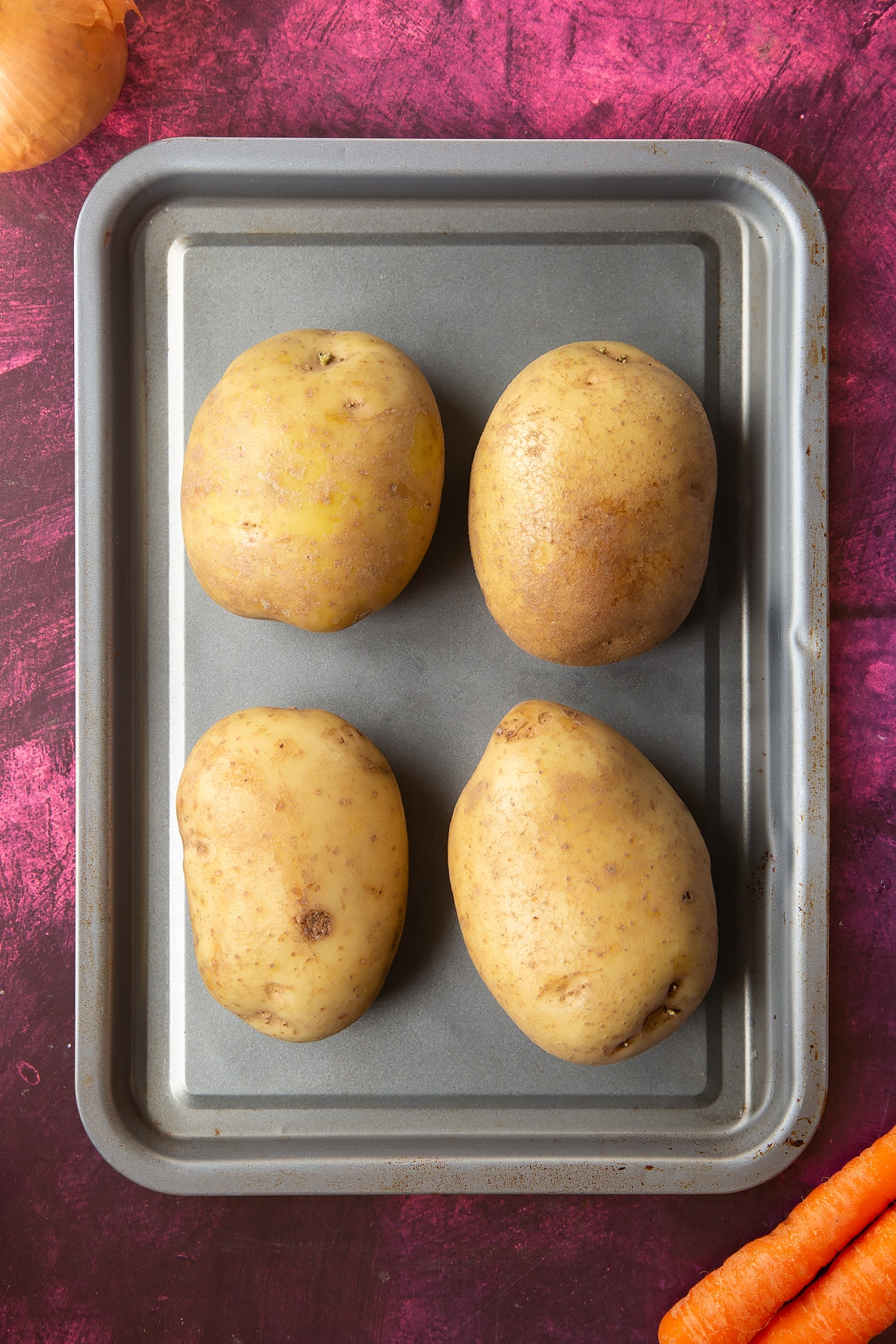 an overhead view of 4 large raw jacket potatos on a silver baking tray