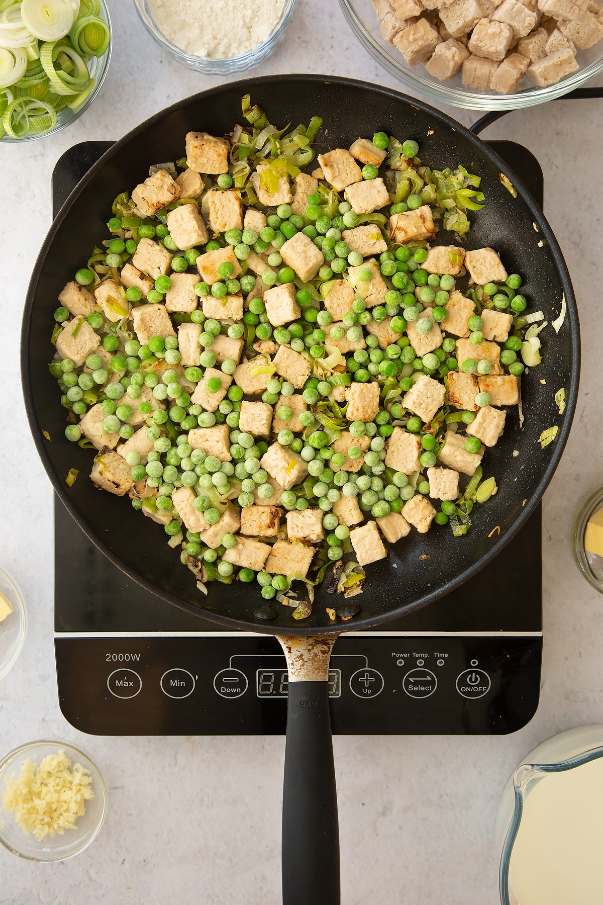 cooked leeks and vegetarian style chicken pieces topped with frozen peas in a large frying pan on an induction hob.