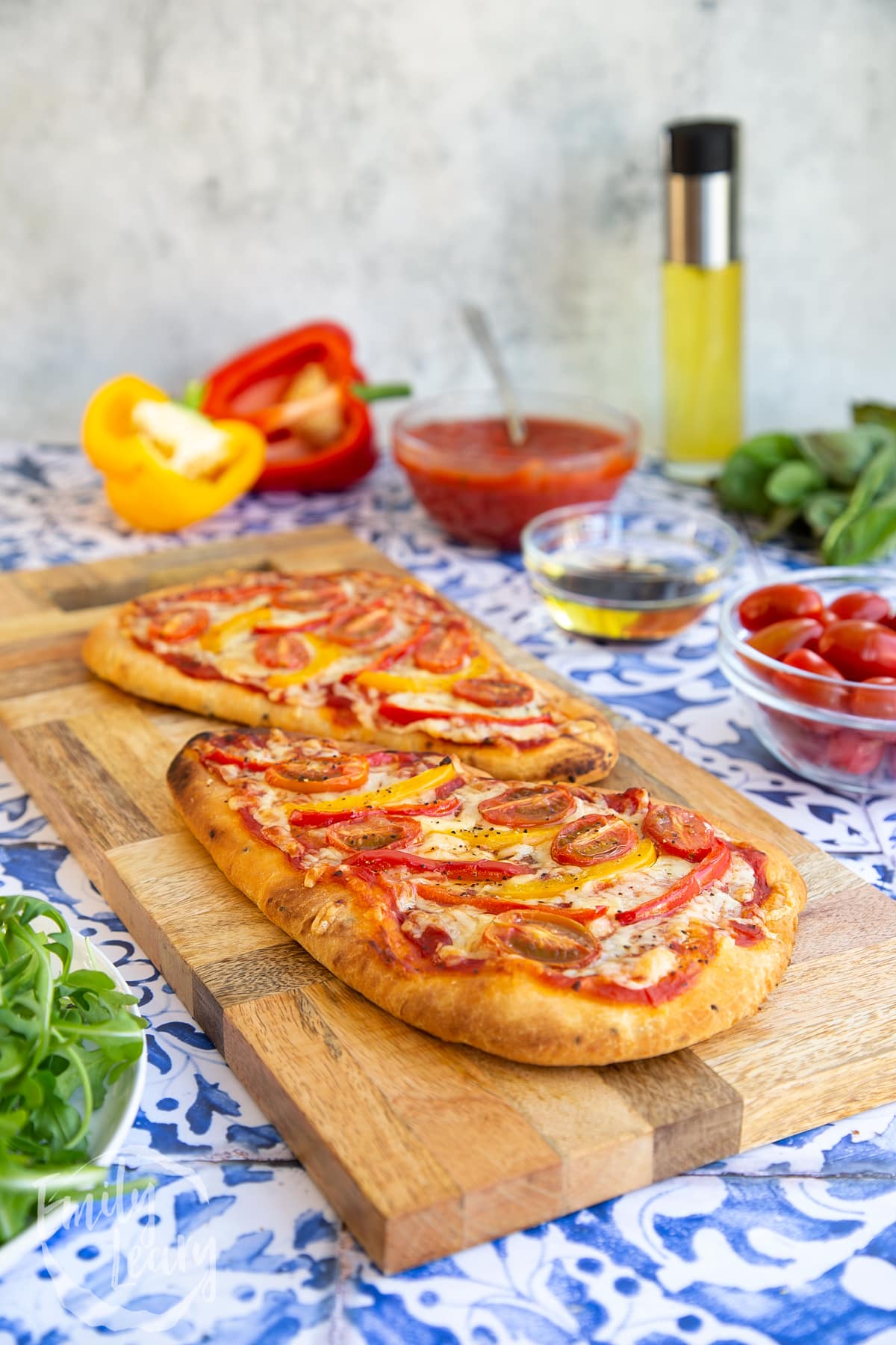 Side on shot of the two Naan bread pizzas on a wooden chopping board which is sat on top of some blue and white decorative tiles. Around the chopping board are some of the ingredients required to make the recipe.