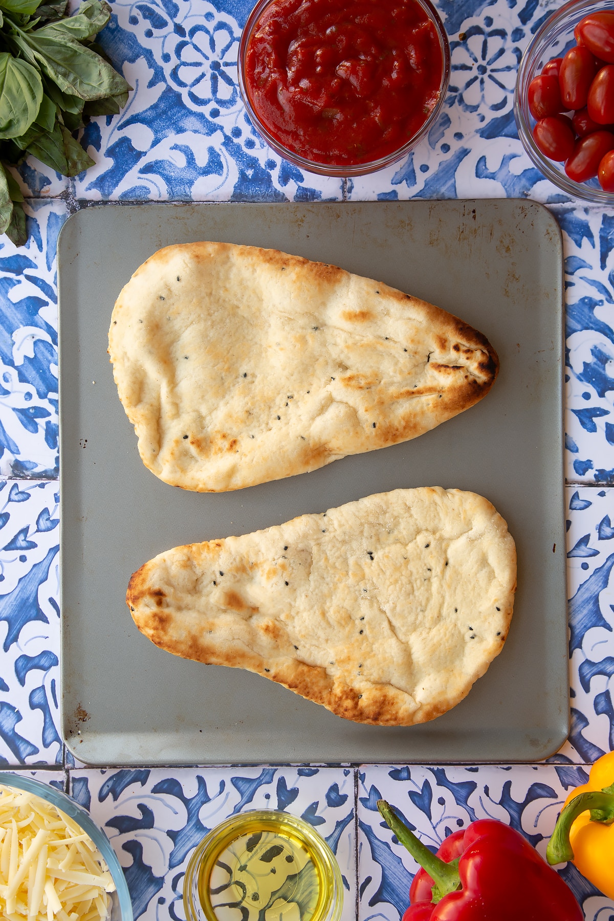 Overhead shot of two plain naan breads on a baking tray with some of the other ingredients required to make the dish surrounding it.