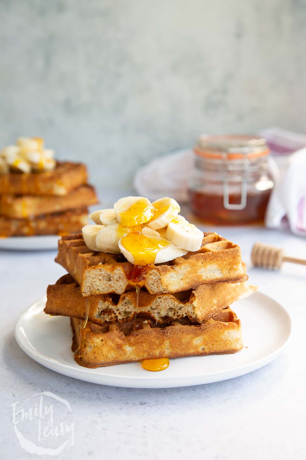 a stack of wholemeal waffles on a white plate topped with honey and banana with a bite missing from the front.