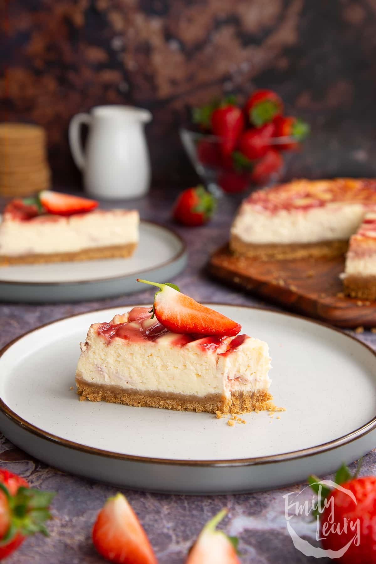 a slice of strawberry swirl cheesecake on a white plate topped with a chopped sttrawberry with a
bite off the front.