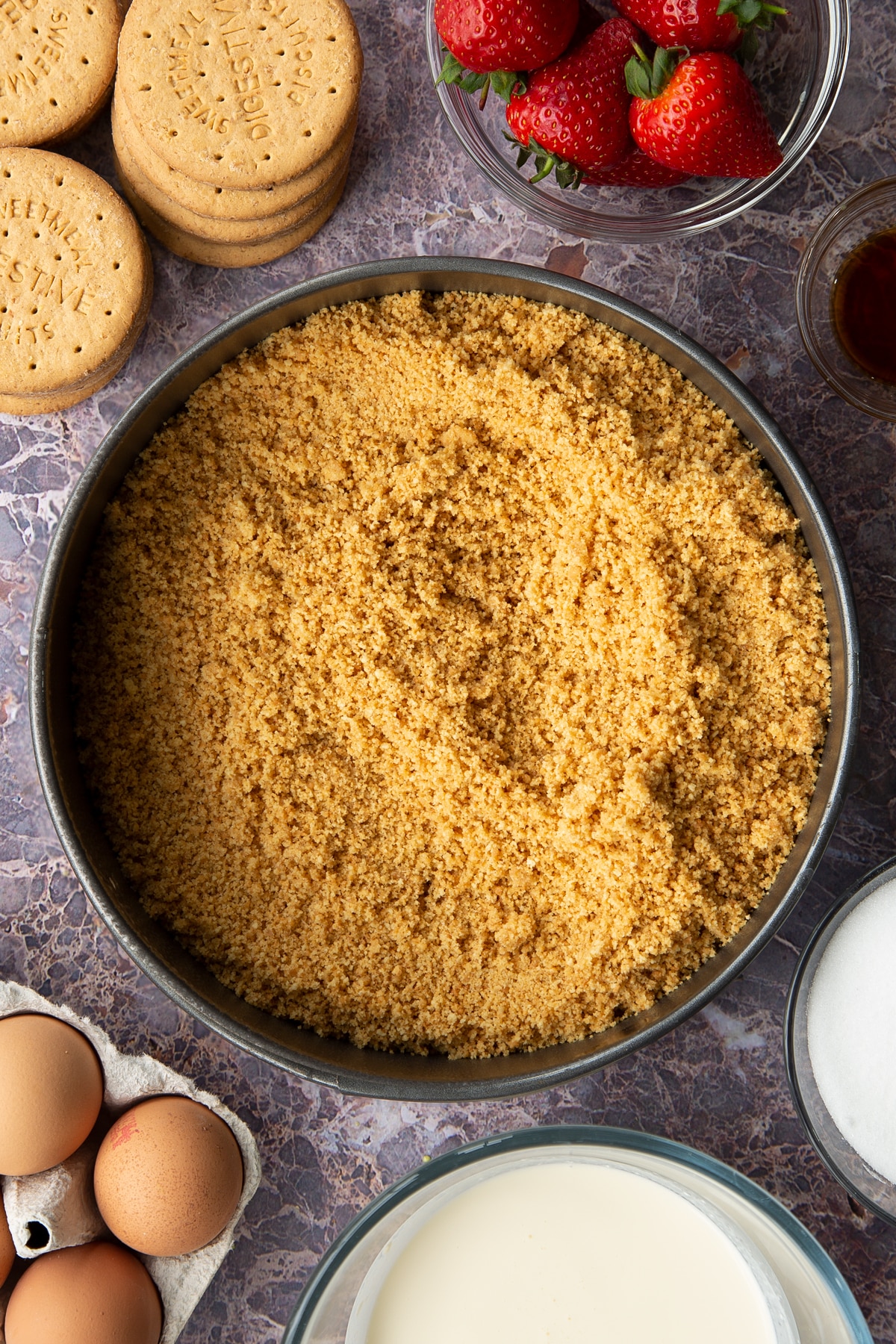 buttery digestive crumbs in a large springform cake tin surrounded by strawberries and digestive biscuits.